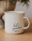 Best Grandma Ever cream campfire mug with black script lettering on a wooden surface, with greenery in the background.