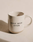 Best Mom Ever cream speckled stoneware mug with black lettering and a natural tan base, photographed in sunlight on a beige surface.