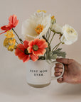 Hand holding a Best Mom Ever cream speckled stoneware mug filled with coral, white, and yellow flowers.
