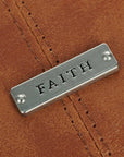 Close-up of Faith metal badge on honey brown faux leather Bible cover showing engraved Faith message and stitched leather detail, Christian Bible cover design