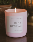 Pink Happy Birthday candle with Tahitian vanilla and buttercream scent burning on a table in a cozy home setting