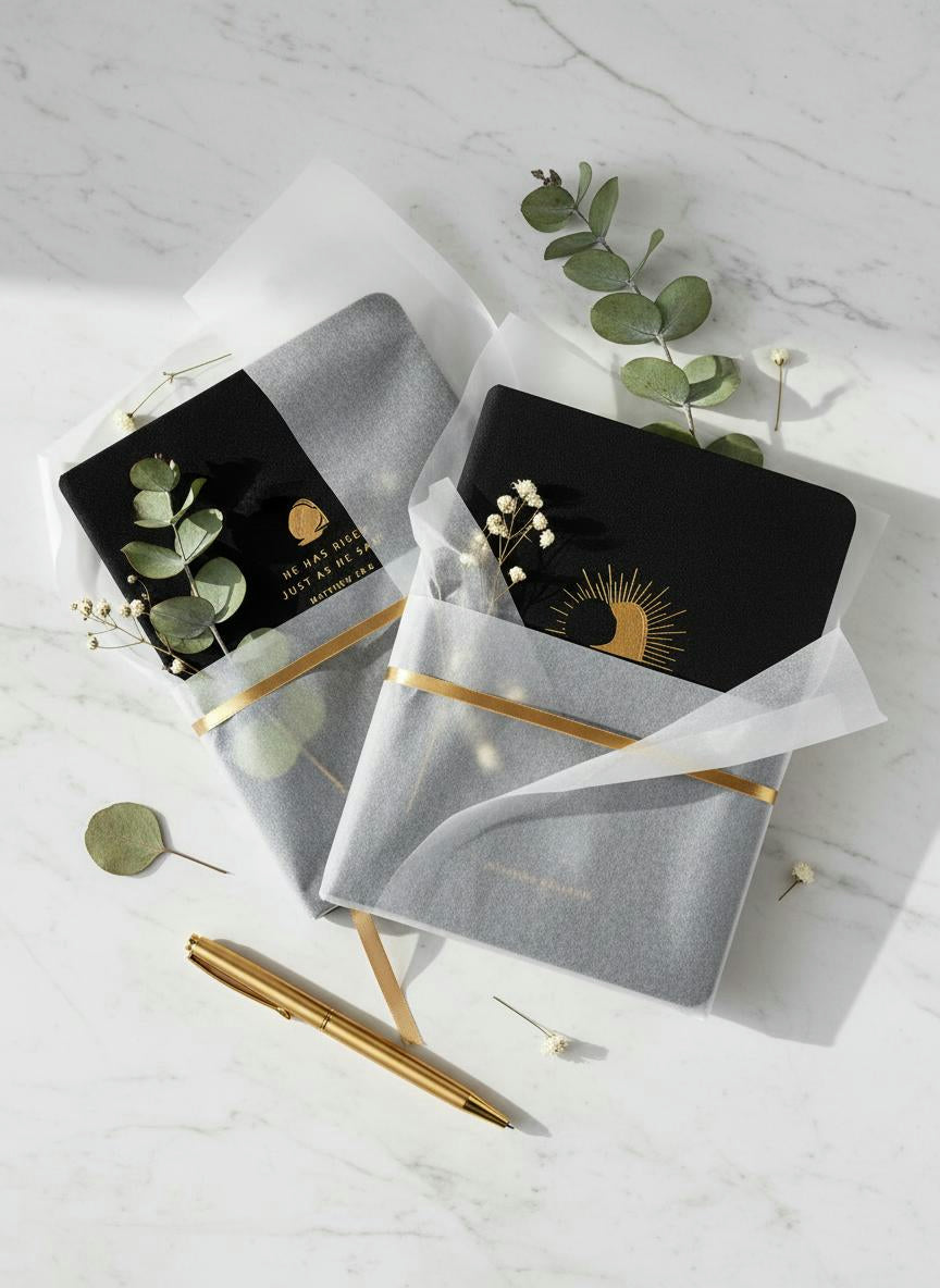 Gift-wrapped Jerusalem Bible and matching journal with gold sunrise artwork, wrapped in tissue paper with greenery on a marble surface.