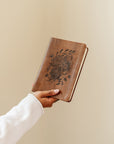 Aurora Christian notebook journal with floral embossed design on warm brown leather-like cover held in hand for prayer journaling, Scripture reflection, and daily devotion