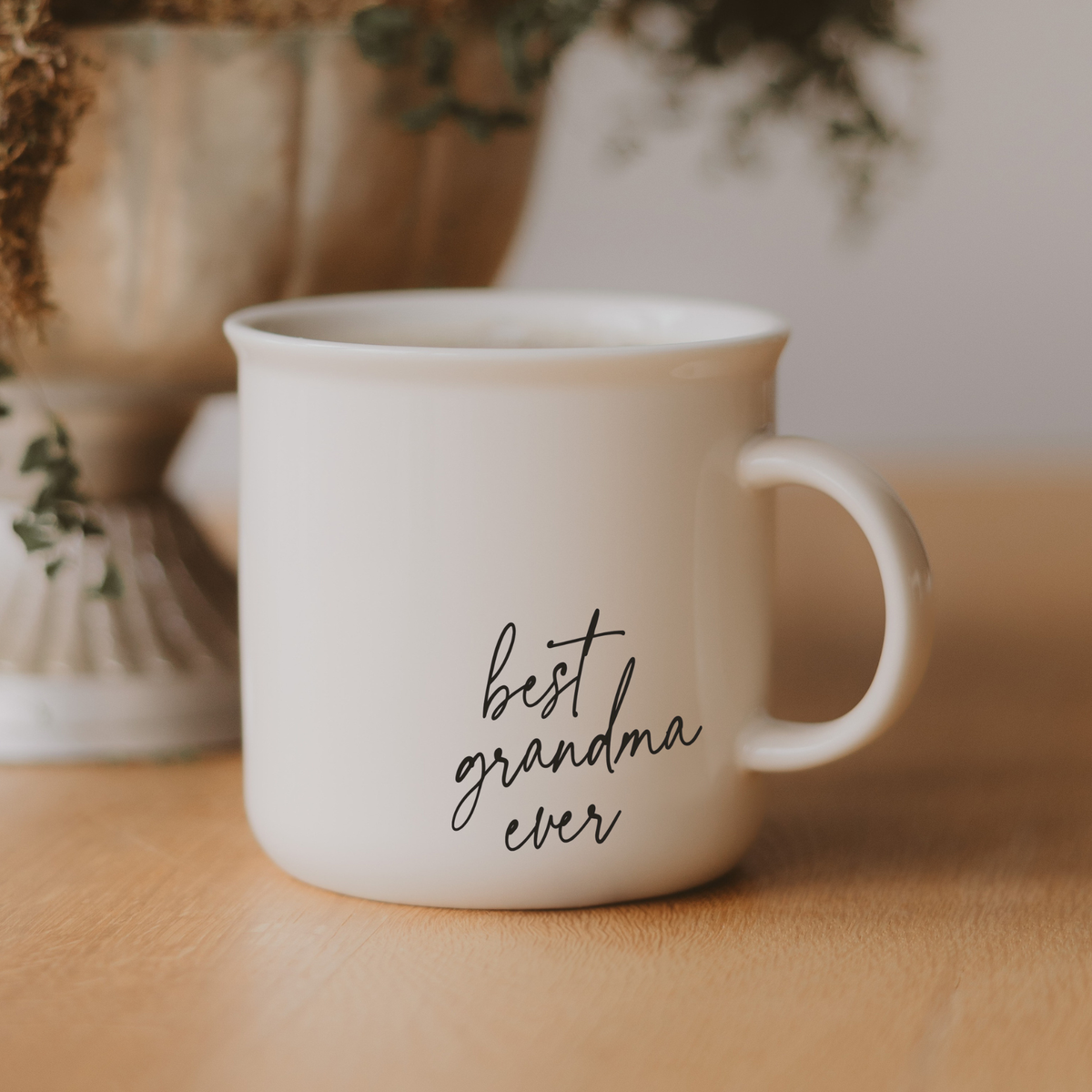 Best Grandma Ever cream campfire mug with black script lettering on a wooden surface, with greenery in the background.