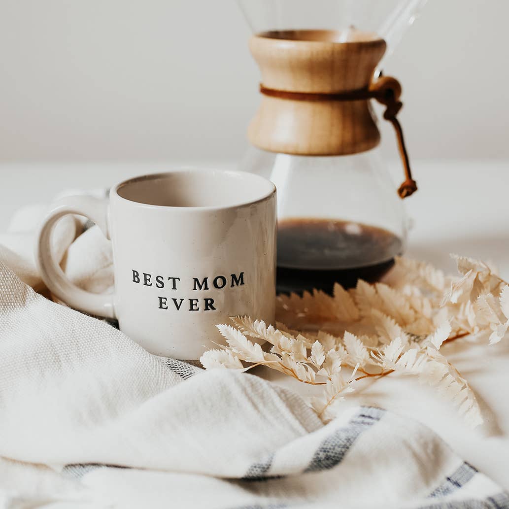 Best Mom Ever cream stoneware mug beside a glass pour-over coffee maker, styled with dried foliage and a white striped cloth.