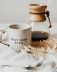 Best Mom Ever cream stoneware mug beside a glass pour-over coffee maker, styled with dried foliage and a white striped cloth.