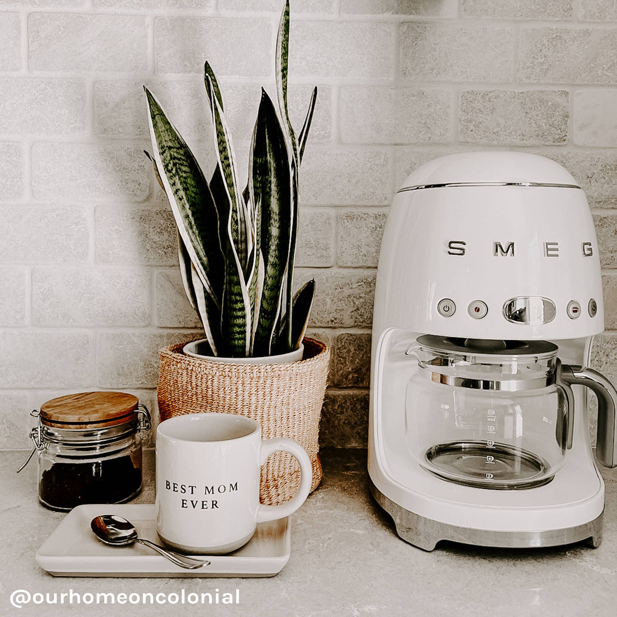 Best Mom Ever cream stoneware mug on a tray with a spoon beside a white coffee maker, coffee jar, and potted snake plant.
