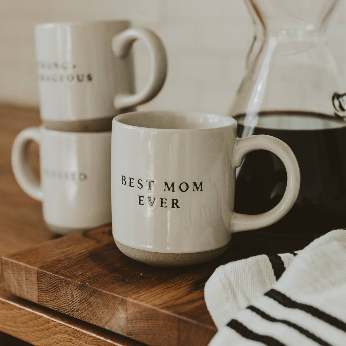 Best Mom Ever cream speckled stoneware mug on a wooden board beside a glass coffee carafe, with stacked mugs in the background.