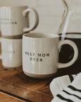 Best Mom Ever cream speckled stoneware mug on a wooden board beside a glass coffee carafe, with stacked mugs in the background.