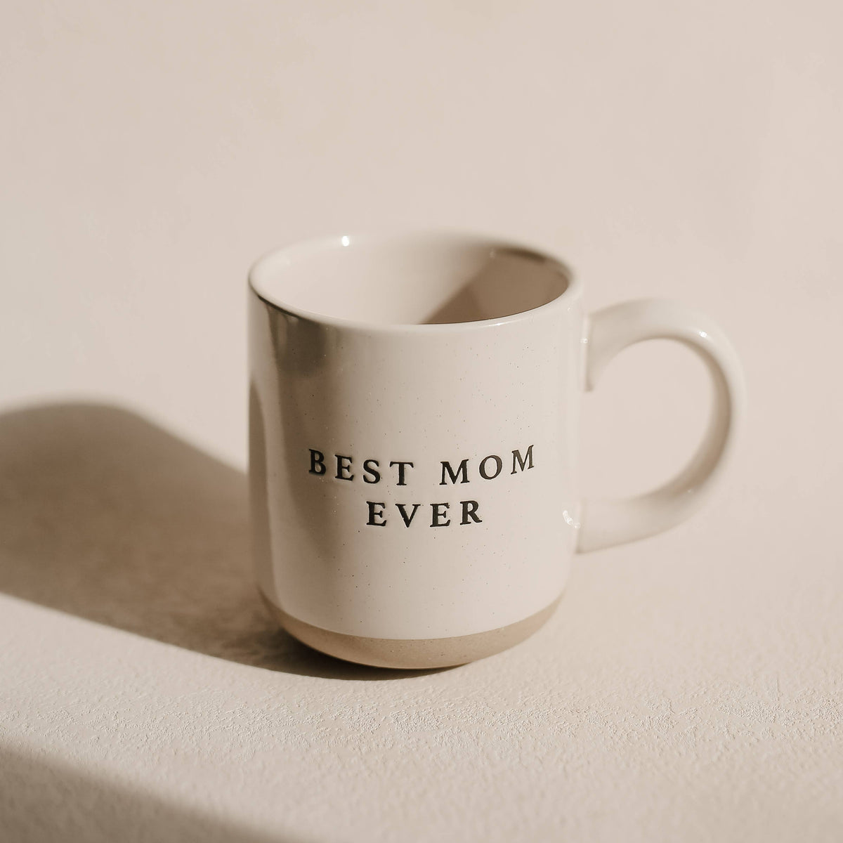 Best Mom Ever cream speckled stoneware mug with black lettering and a natural tan base, photographed in sunlight on a beige surface.