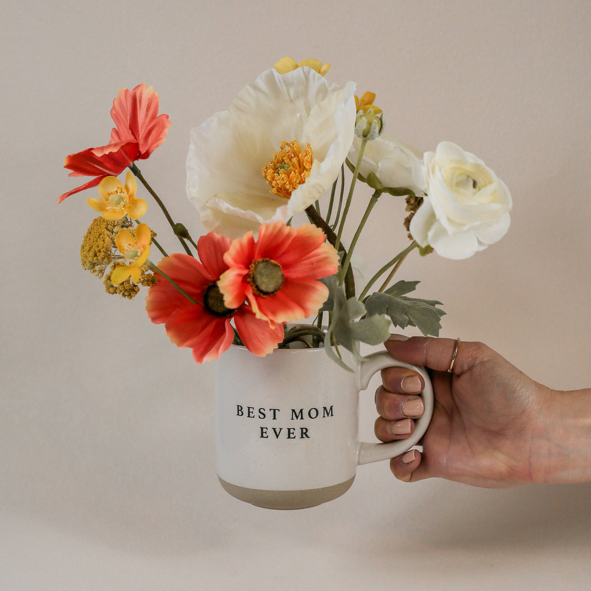 Hand holding a Best Mom Ever cream speckled stoneware mug filled with coral, white, and yellow flowers.
