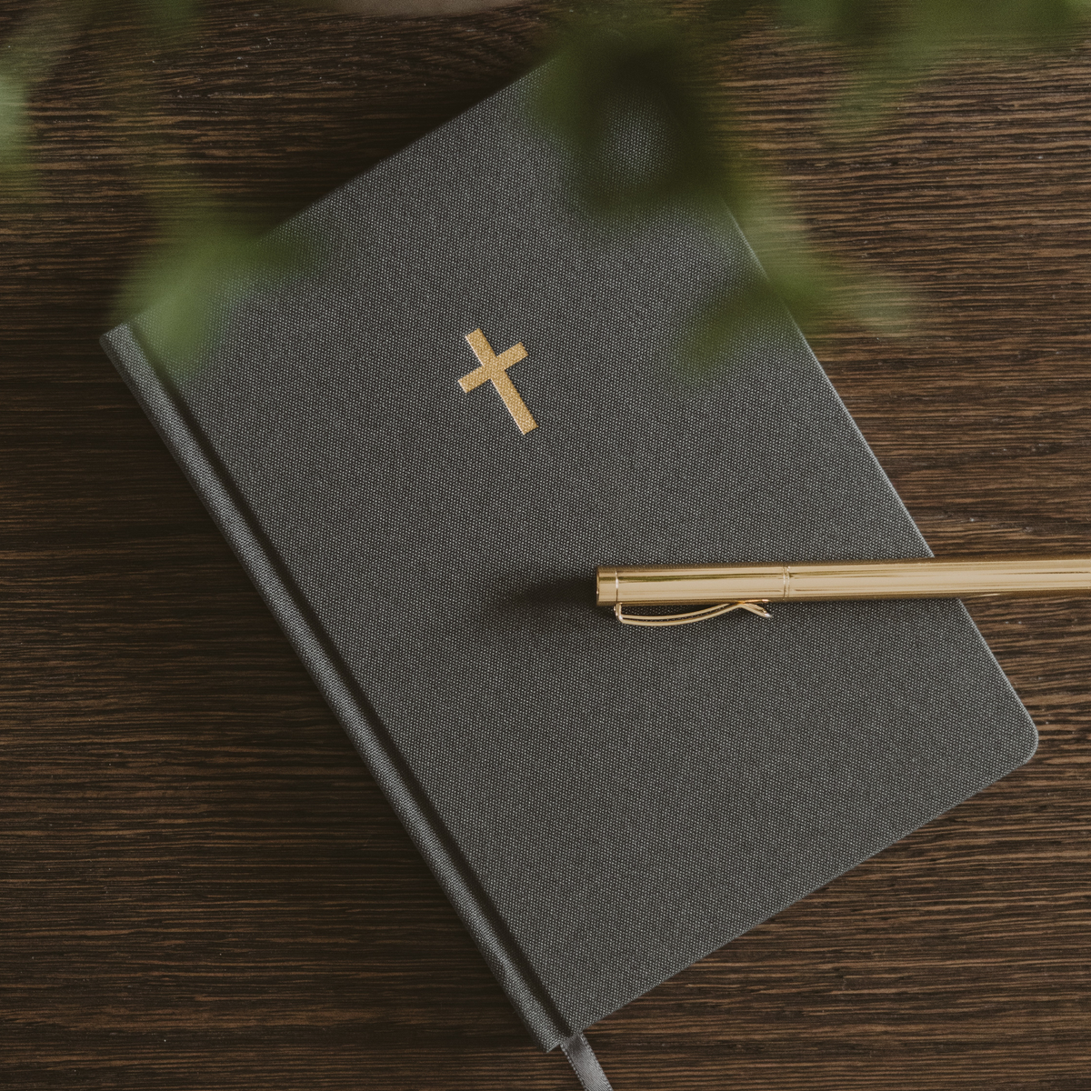 Grey fabric hardcover sermon notes journal with a gold foil cross and grey ribbon bookmark, styled with a gold pen on a wooden table.