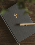 Grey fabric hardcover sermon notes journal with a gold foil cross and grey ribbon bookmark, styled with a gold pen on a wooden table.