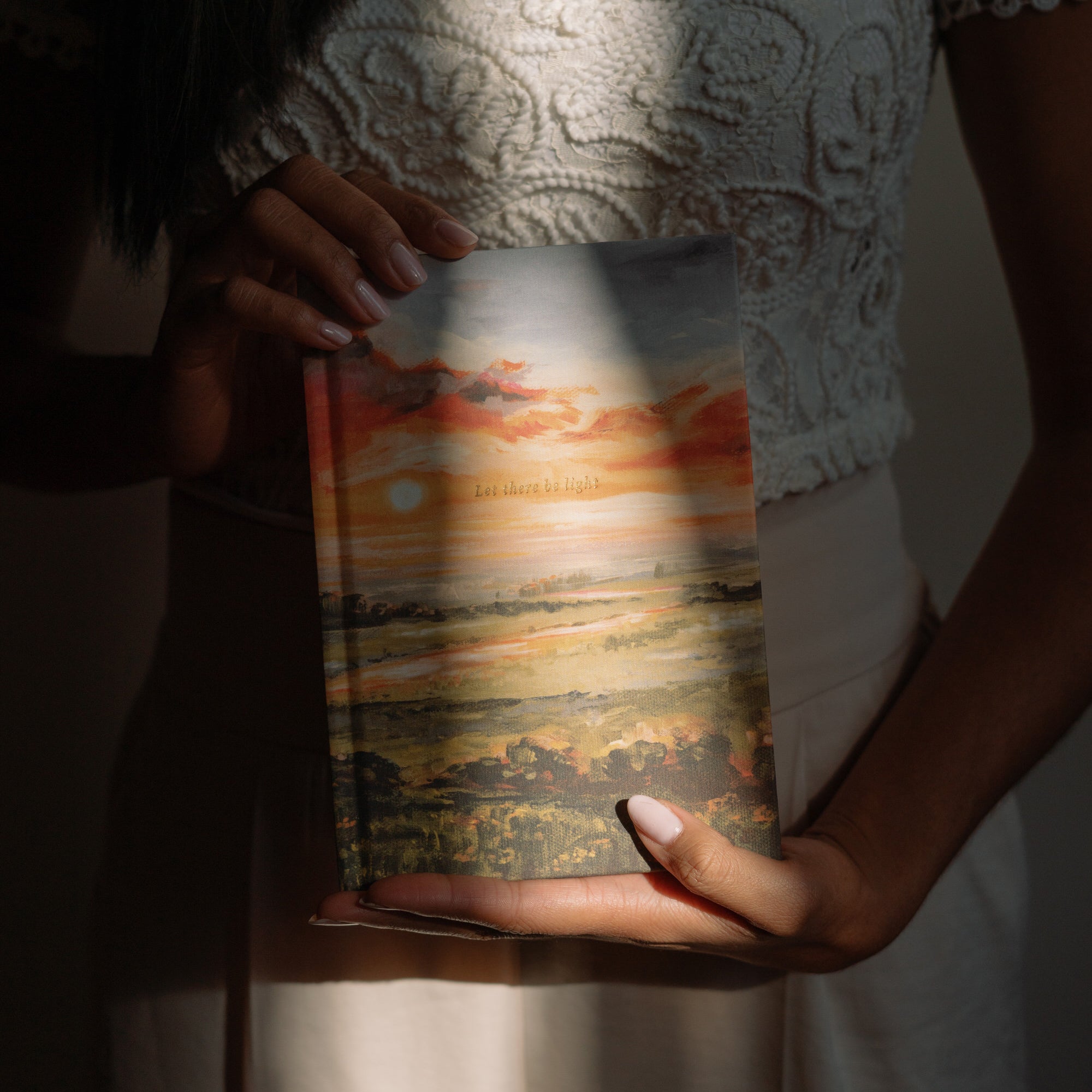 Hands holding Florence Christian notebook with Let there be light sunset cover design in warm natural light