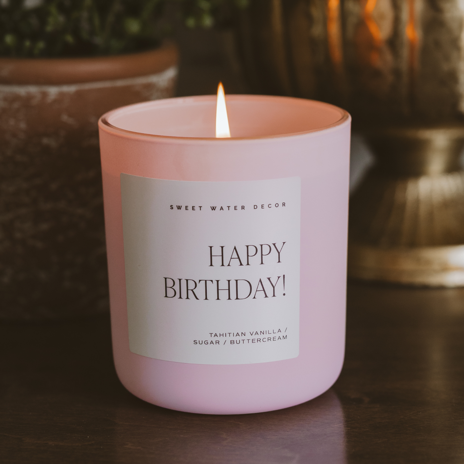 Pink Happy Birthday candle with Tahitian vanilla and buttercream scent burning on a table in a cozy home setting