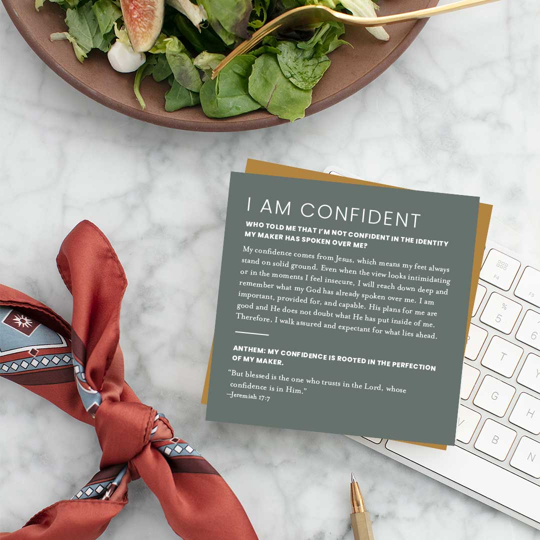 Lifestyle photo of the ‘I Am Confident’ Christian identity affirmation card from the Identity Scripture Cards set, styled on a marble desk beside a salad bowl, pen, keyboard, and scarf—encouraging believers with Jeremiah 17:7 and confidence rooted in Christ.