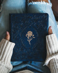 Close-up of gold floral design on Versailles NLT Bible cover inspired by French palace gardens being held by a woman with a cream colored sweater and jeans. 